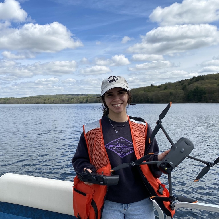 A photo of Jalissa Pirro in an orange safety vest holding a drone at the edge of a body of water